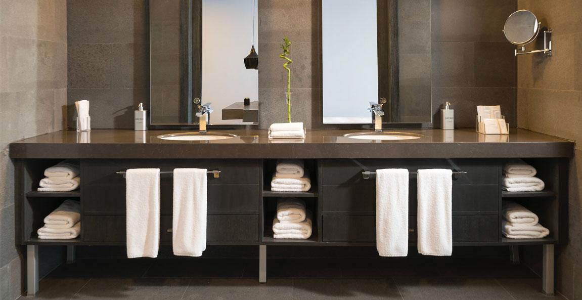 A bathroom countertop and vanity with two sinks and mirrors, hanging and folded towels, and a small bamboo plant in the middle showcasing the need for regular bathroom vanity repairs and luxury hotel furniture touch-up